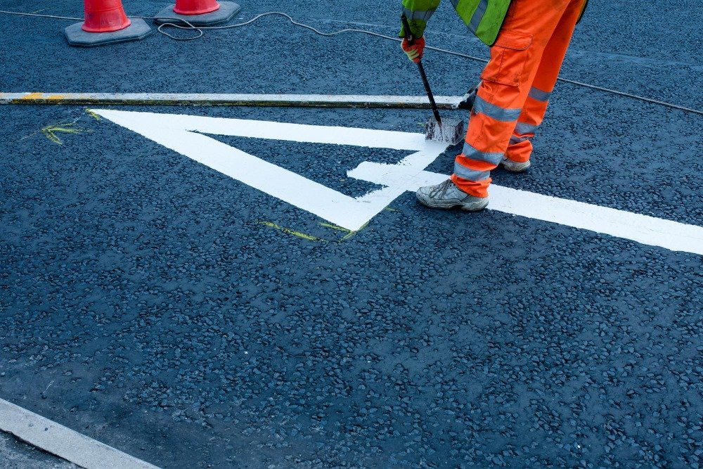 Worker In Orange High-Visibility Clothing Painting White Road Markings On Freshly Paved Asphalt Using A Brush And Guide