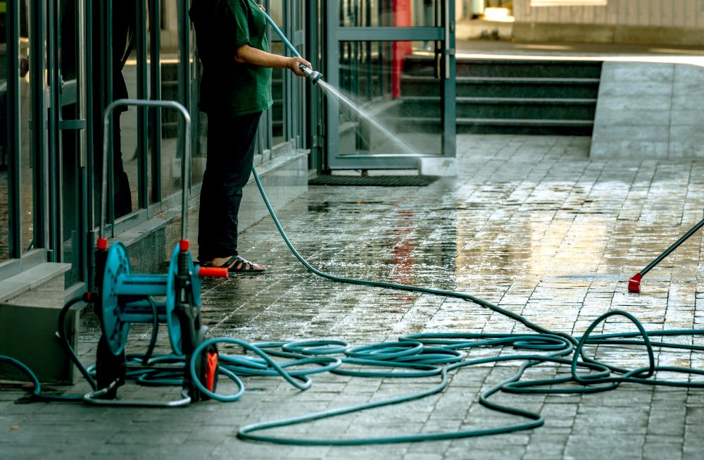 Person Using A High-Pressure Hose To Clean A Wet Sidewalk Outside A Building